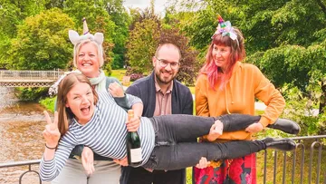 Three people hold a woman horizontally, outdoors.