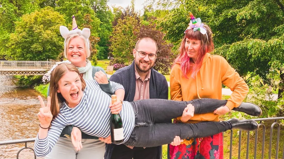 Three people hold a woman horizontally, outdoors.