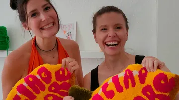 Two women holding colorful handmade rugs indoors.