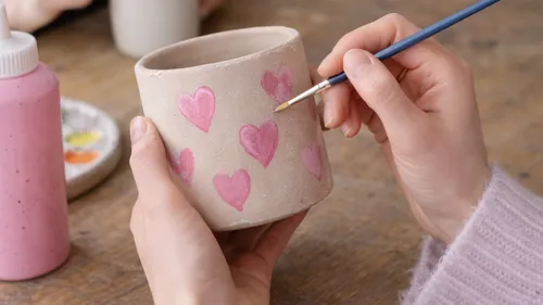 Hands painting pink hearts on ceramic mug.