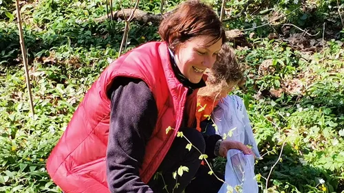 Person in red vest smiling, picking plants in forest.