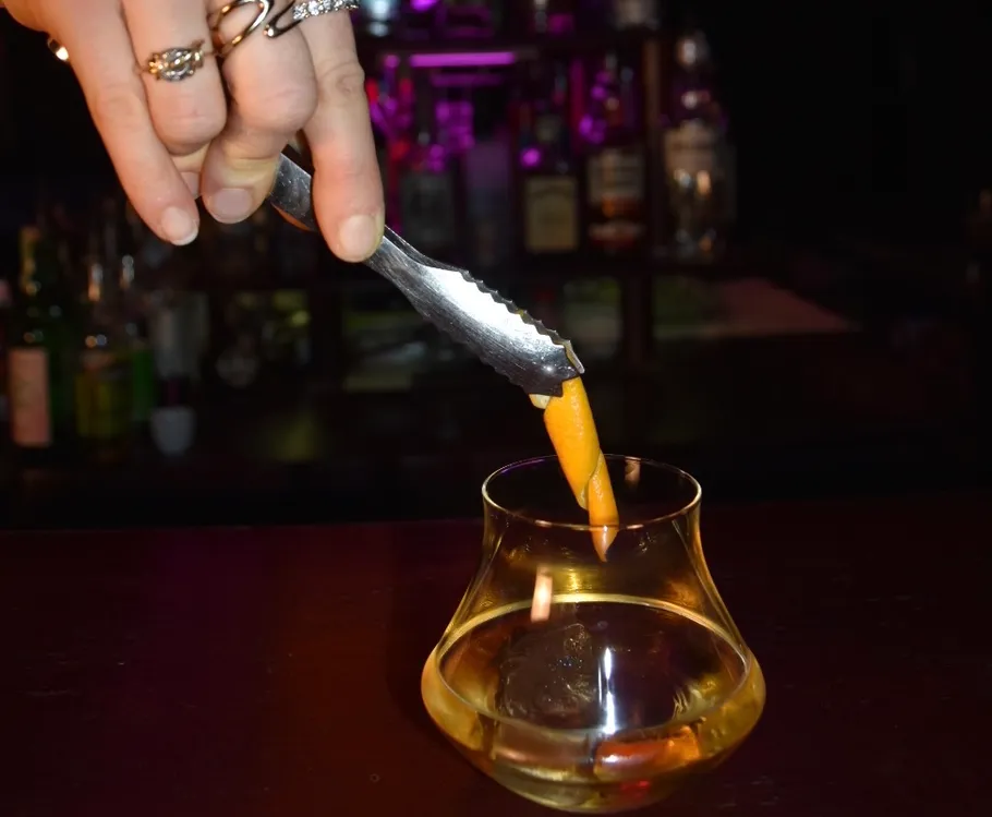 Hand adding orange peel to cocktail glass.
