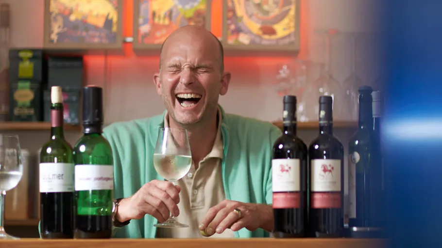 Man laughing, holding wine glass, surrounded by bottles.