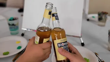 Two hands clinking Bionade bottles at a table.