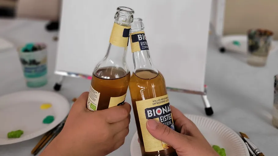 Zwei Hände stoßen Bionade-Flaschen am Tisch an.