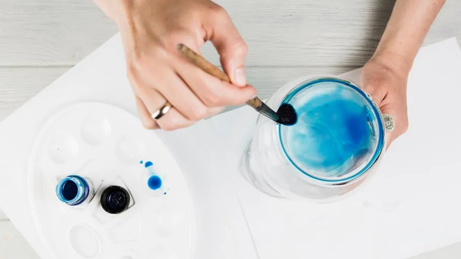 Person painting glass jar with blue paint.