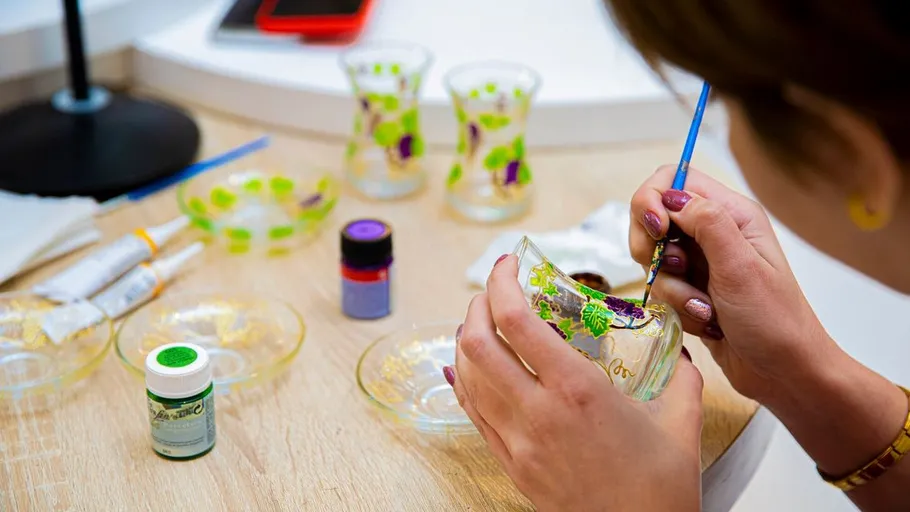 Person painting designs on glass cups, tabletop setting.
