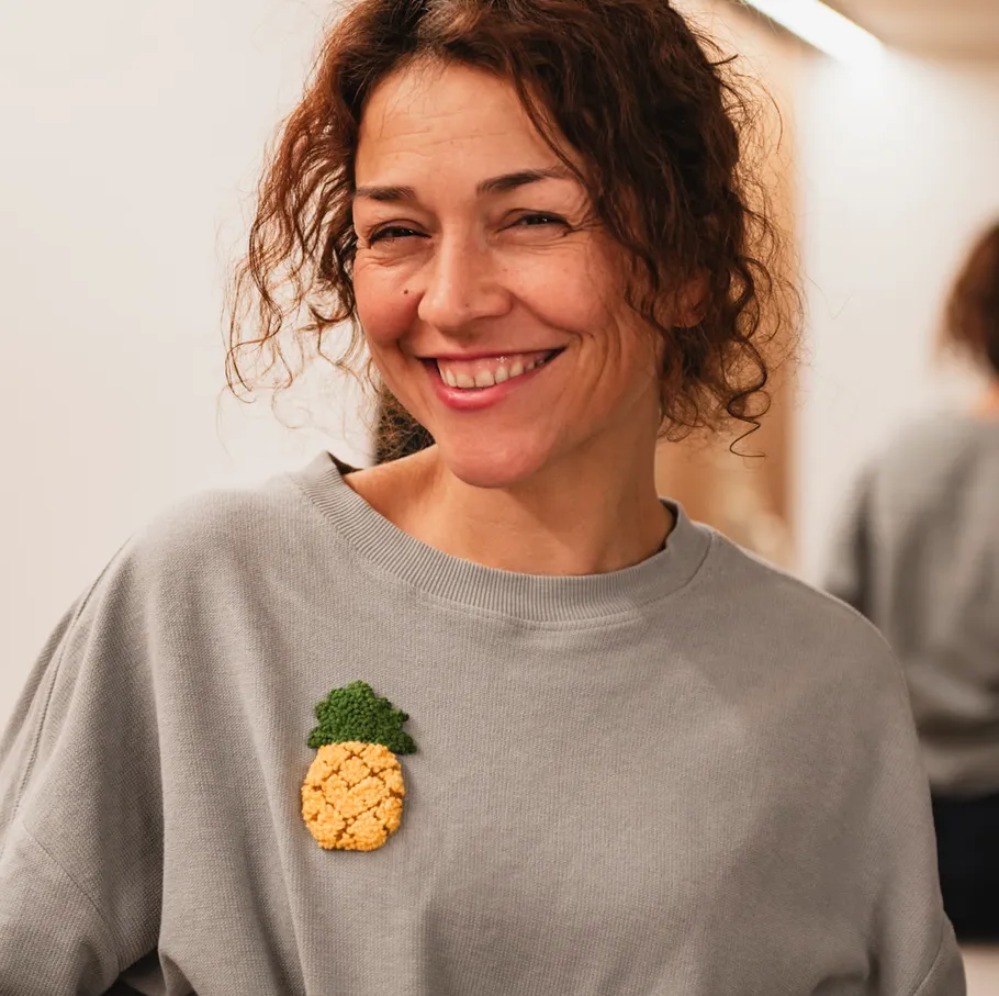 Lächelnde Frau mit grauem Pullover und Ananas-Aufnäher.