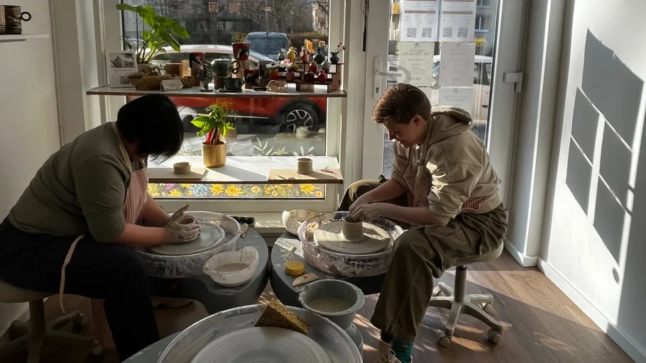 Two people crafting pottery indoors by window.