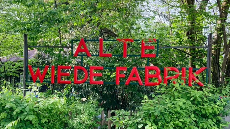 Red sign saying 'Alte Wiede Fabrik' among trees.
