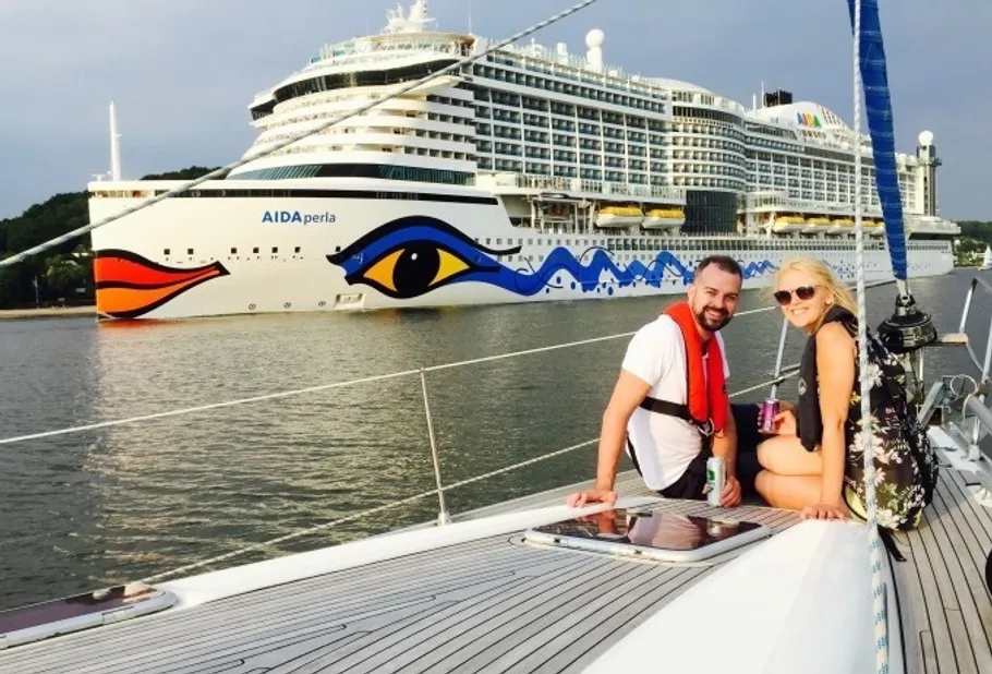 Two people smiling on sailboat near cruise ship.