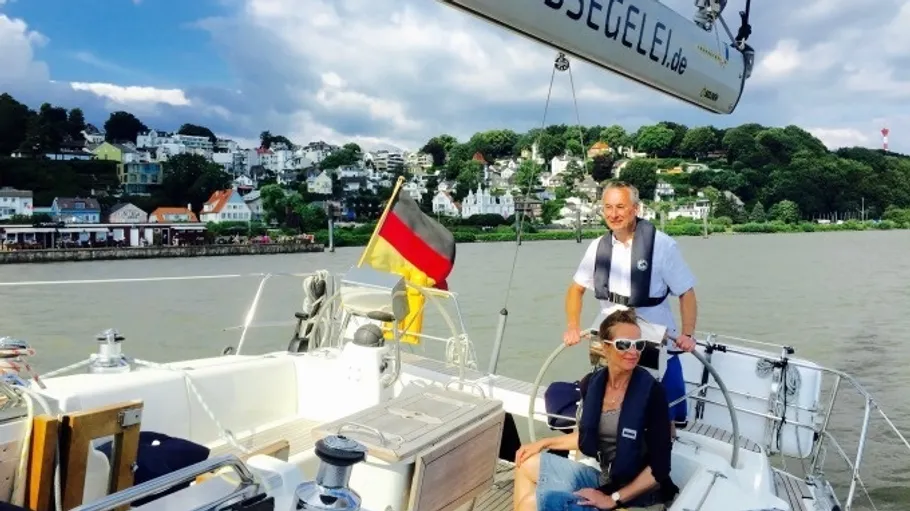 Zwei Personen segeln auf Boot; Küstenstadt im Hintergrund.