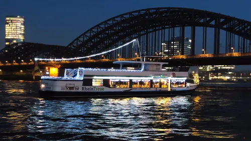 Beleuchtetes Boot fährt nachts unter Kölner Brücke.