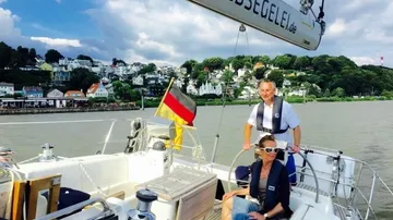 Zwei Personen segeln auf Fluss, Stadt dahinter.