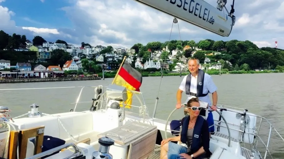 Two people sailing boat on river, town behind.