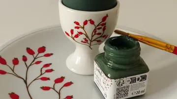 Eierbecher mit grünem Ei, Farbdose und Pinsel.