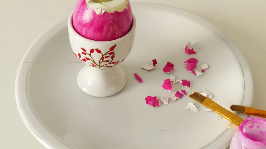 Bemaltes Ei im Becher mit rosa Schalenstücken.