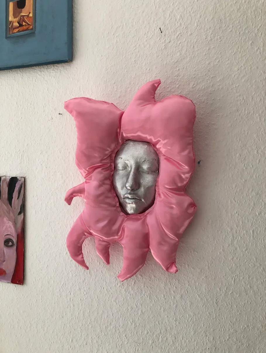 Silver face in pink fabric frame on wall.