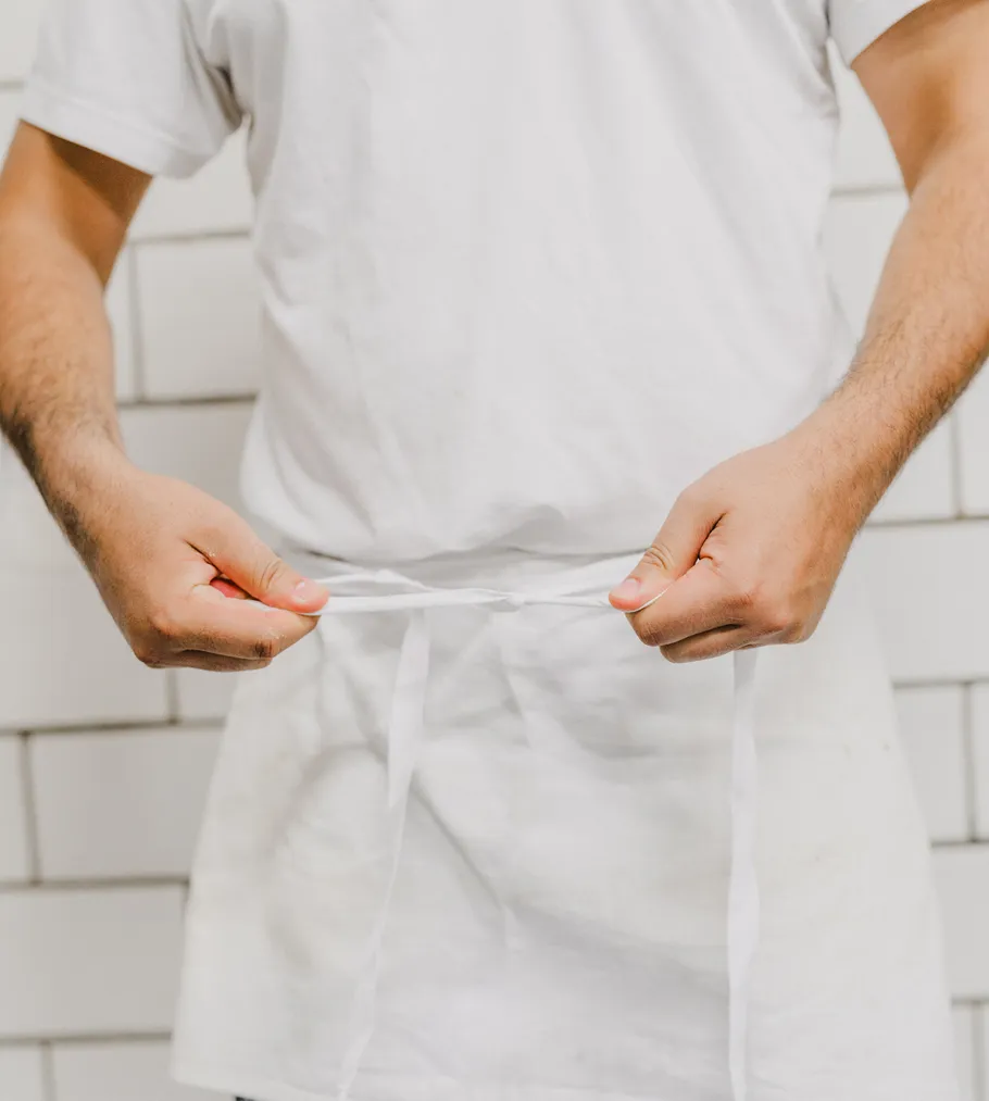 Person tying white apron against tiled wall.