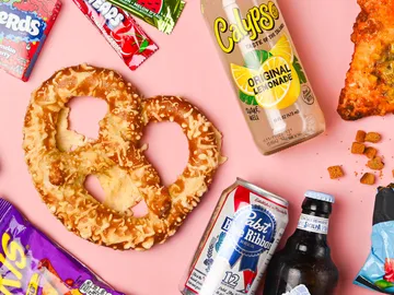 Brezel liegt auf pinkem Hintergrund, umgeben von Snacks und Getränken.