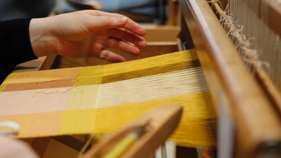Hands weaving yellow fabric on a loom.
