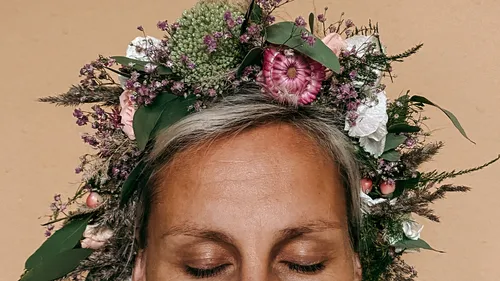 Person trägt Blumenkrone, Augen zu, beiger Hintergrund.