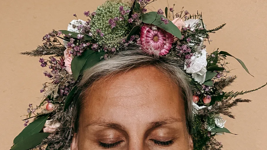 Person trägt Blumenkrone, Augen zu, beiger Hintergrund.