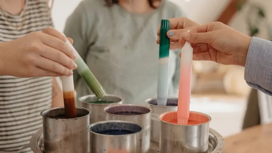Hands dipping candles into colorful wax pots.