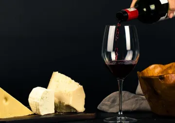 Red wine is poured into a glass over cheese and wooden bowl.