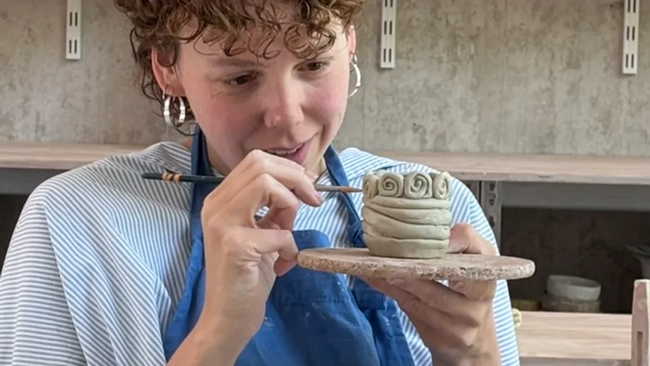 Person modelliert Tonkeramik drinnen, lächelnd.