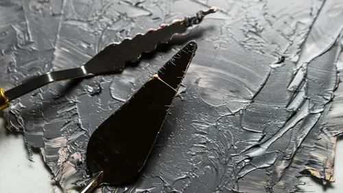 Palette knives spreading dark paint on canvas.