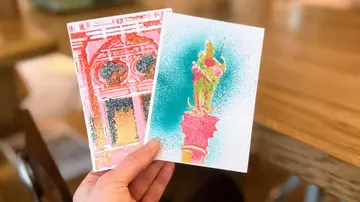 Hand hält zwei bunte Postkarten in Innenraum.