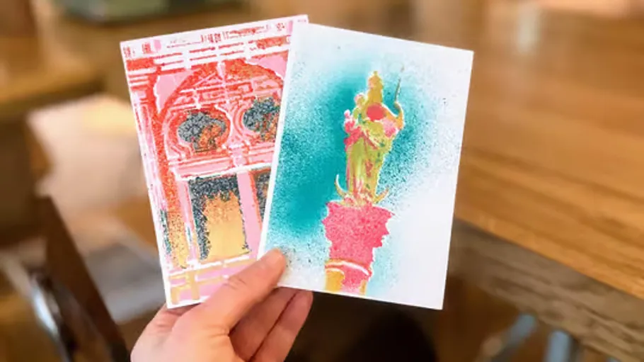 Hand hält zwei bunte Postkarten in Innenraum.