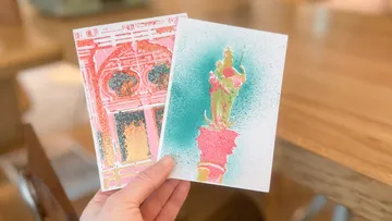 Hand hält zwei bunte künstlerische Postkarten drinnen.