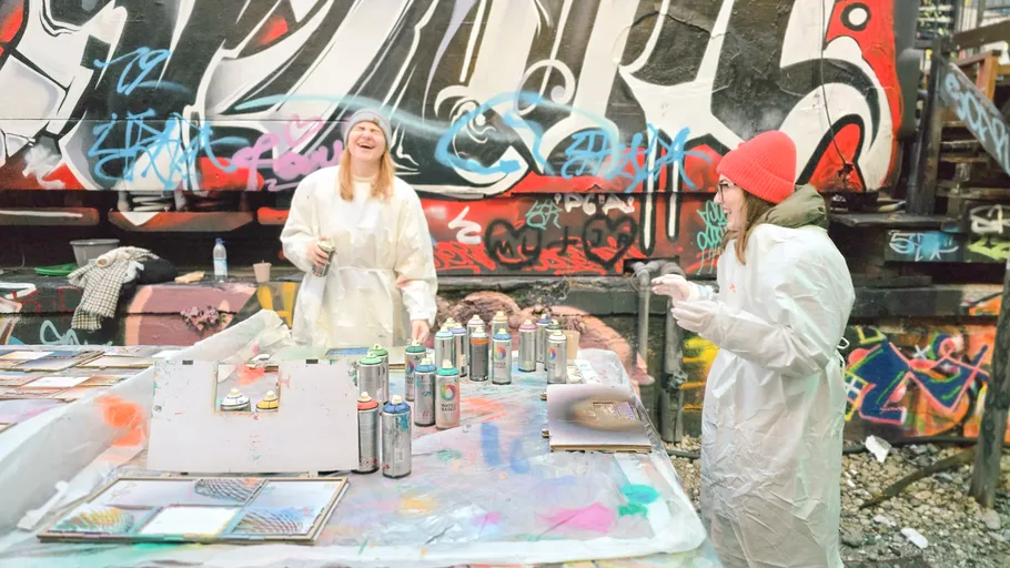 Zwei Personen sprühen draußen, Graffiti im Hintergrund.