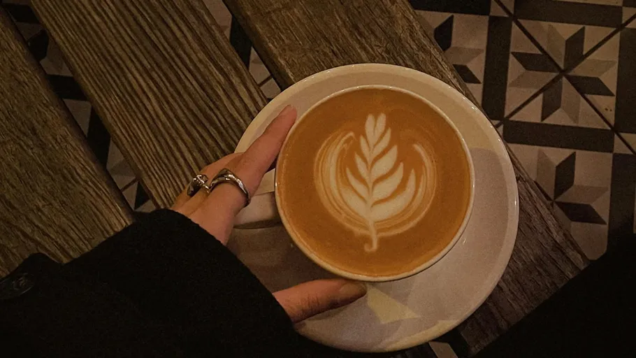 Hand holding latte with leaf art, wooden table.