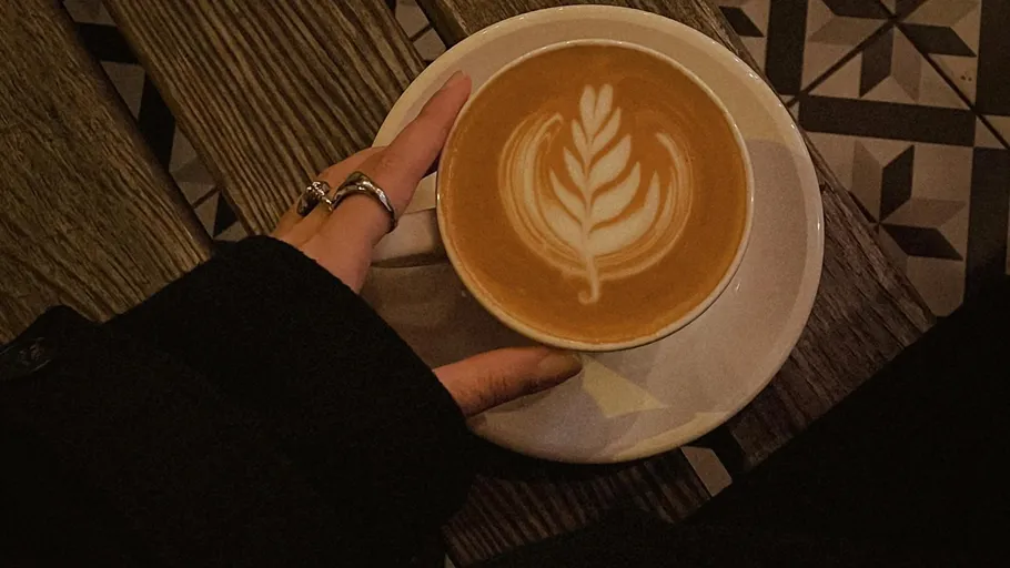 Hand hält Latte mit Blattkunst auf Tisch.
