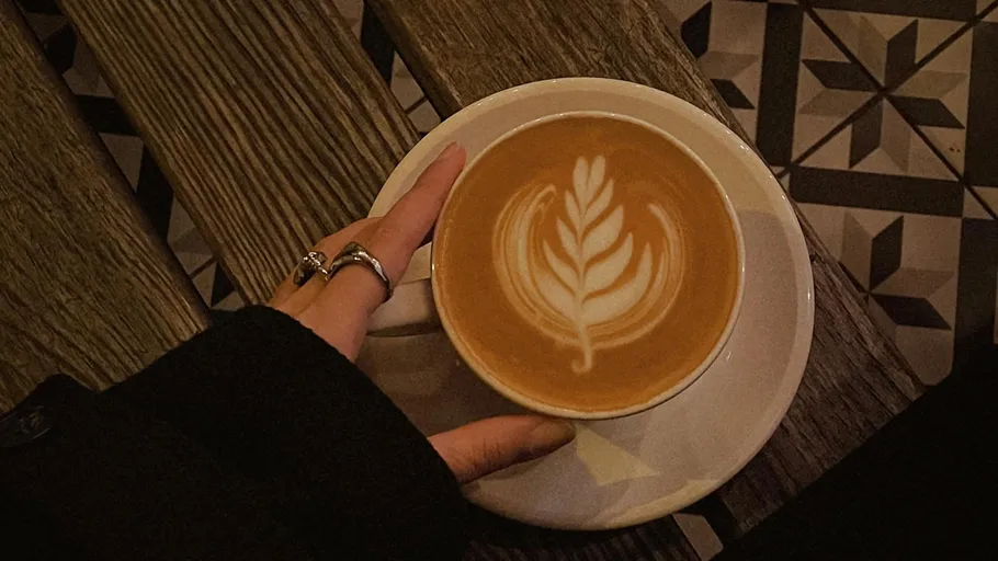 Hand holds latte with leaf art on bench.