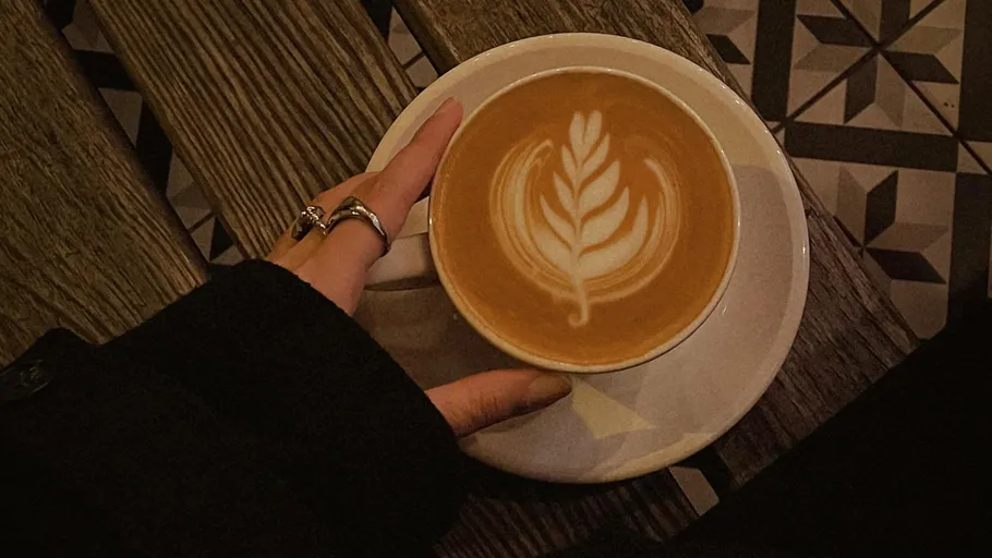 Hand hält Latte mit Blätterkunst auf Tisch.
