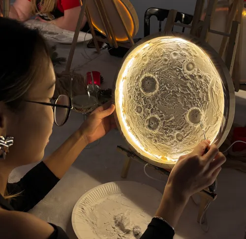Woman sculpting illuminated moon model at table.