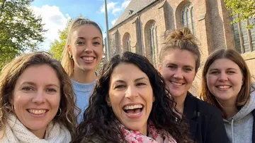 Five smiling women outside near a building.