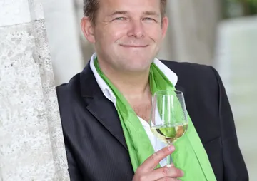 Mann mit Weinglas lächelt vor einer hellen Kulisse.