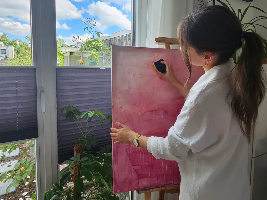 Frau malt pinke Leinwand drinnen am Fenster.