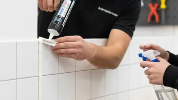 Person trägt Dichtmittel auf weiße Fliesenwand auf.