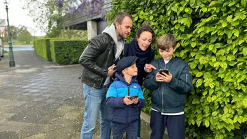 Familie schaut auf Handys nahe grüner Hecke.