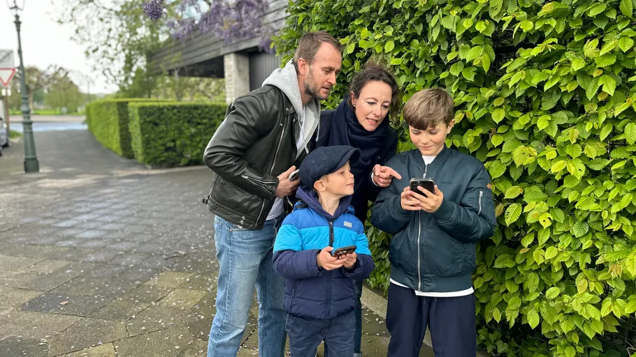 Familie draußen nutzt Smartphones nahe Büschen.