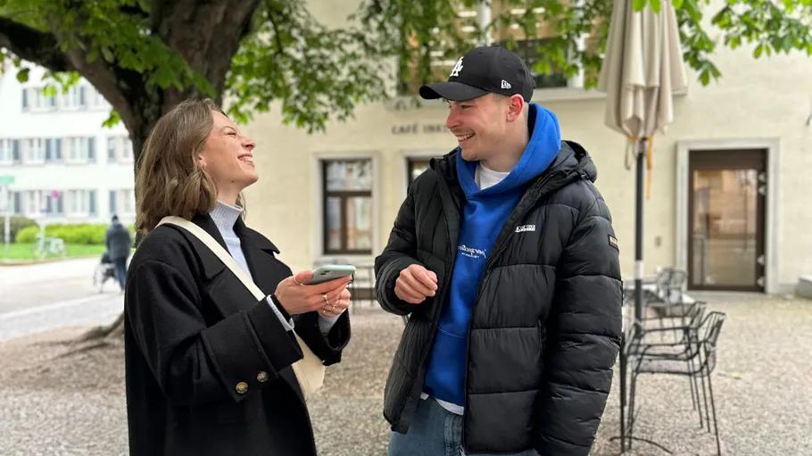 Two people smiling and talking outside, under a tree.