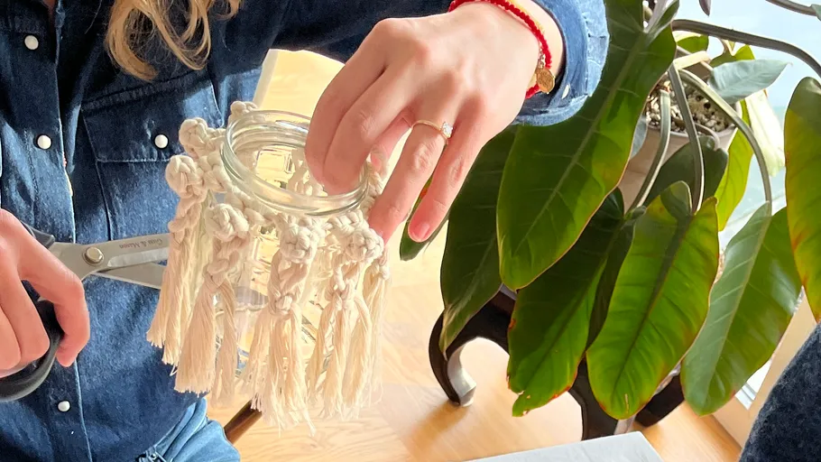 Person bastelt Makramee-Glas neben Pflanze.
