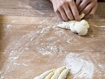 Hände formen Teig auf einer bemehlten Holzplatte.