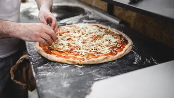 Person fügt Belag zu Pizza in Küche hinzu.
